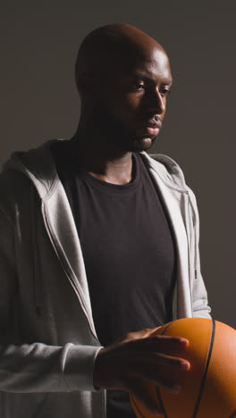 Vertical-Video-Studio-Vertical-Video-Portrait-Shot-Of-Male-Basketball-Player-Wearing-Tracksuit-Holding-Ball-Against-Dark-Background